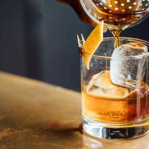 A cocktail being poured into a glass with ice and a citrus twist garnish, set on a bar counter.